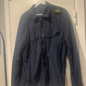 Marinblå vindjacka / overshirt från Stone Island - Säljer en marinblå vindjacka/overshirt från Stone Island med klassisk patch på axeln. Jackan har dragkedja framtill, två bröstfickor med lock och långa ärmar. Materialet är lätt och syntetiskt, perfekt för blåsiga dagar. Stilsäker och enkel att matcha med jeans eller cargo.