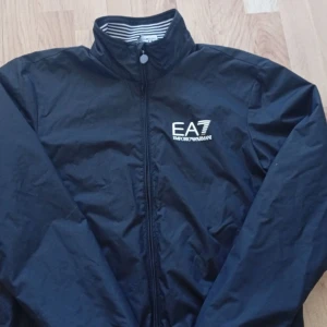   EA7 Emporio Armani Jacka perfekt till Höst XS - Svart vindjacka från EA7 Emporio Armani med vitt logotryck på bröstet. Jackan har dragkedja, ståkrage och coola vita ränder på insidan av kragen samt vid ärmslut och midja. Tillverkad i lätt polyester, perfekt för  och höst Väder Storlek XS men passar S också .kommer från djur och rökfritt hem . Finns ute på andra sidor med 