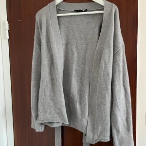 Grå kofta med knytband från Lager 157 - Mysig grå kofta från Lager 157 i stickad bomullsmix. Modellen är öppen framtill och har långa ärmar samt knytband i midjan för en snygg passform. Perfekt att slänga över en t-shirt eller linne när det är lite kyligare ute.