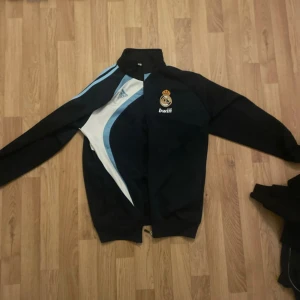 Real Madrid adidas zip tröja svart/blå - Säljer en svart och blå vindjacka från adidas med Real Madrid-logga och Bwin-tryck. Jackan har dragkedja framtill, vita och ljusblå detaljer samt lång ärm. Perfekt för dig som vill ha sportig och clean stil.