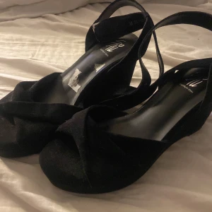 Svarta kilklack sandaler från din sko - Säljer ett par svarta sandaler med kilklack från H&M. Skorna har breda korslagda remmar över tårna och en justerbar rem runt ankeln. Materialet är mocka-liknande syntet och sulan är platt framtill med hög kilklack. Perfekta för sommarkvällar.
