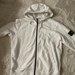 Vit hoodie från Stone Island - Vit hoodie från Stone Island med klassisk svart och gul logotyp-patch på vänster ärm. Tröjan har huva med snörning, hel dragkedja framtill och två stora fickor. Tillverkad i mjuk bomull, perfekt för chill dagar. Snygg och enkel design som funkar året runt. Inte använd på länge. Inga skador eller liknade. Först till kvarn, fraktar alltid samma dag.