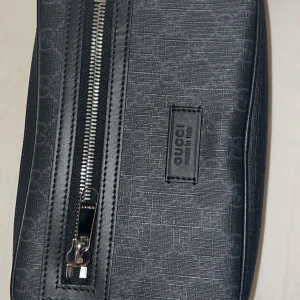 Svart Gucci axelväska med monogram - Snygg svart axelväska från Gucci med diskret GG-monogram över hela väskan. Väskan har en silverfärgad dragkedja framtill och bred justerbar axelrem med röd-blå rand. Tillverkad i skinn och textil, perfekt för dig som vill ha en stilren och lyxig accessoar.