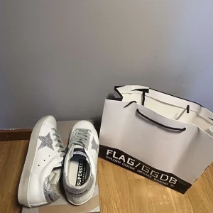 Golden Goose Superstar sneakers silver - Golden Goose Superstar sneakers i vitt skinn med silverglittrig stjärna på sidan och silverglittrig häl. Klassisk låg modell med snörning och platt sula. Snygga detaljer och ikonisk branding på plösen och insidan. Perfekta för dig som vill sticka ut med stil.