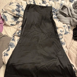 Svart långkjol i satin med spetskant - Snygg svart långkjol i glansigt satinliknande material med en elegant spetskant upptill. Kjolen har en rak och stilren passform som ger en classy vibe. Perfekt för dig som gillar en enkel men ändå chic look.