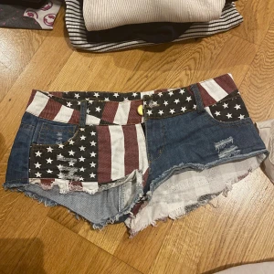 Jeansshorts med USA-flagga och slitningar - Säljer ett par coola jeansshorts med amerikansk flaggdesign i rött, vitt och blått. Shortsen har stjärnor och ränder, slitna detaljer och fransig kant nertill. Perfekta för sommaren och festivaler, riktigt statement-plagg för dig som vill sticka ut.