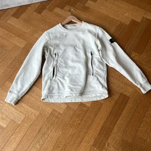 Vit sweatshirt från Stone Island - Vit sweatshirt från Stone Island med klassisk rund hals och två dragkedjeförsedda fickor framtill. Tröjan har lång ärm och den ikoniska loggan på vänster ärm. Perfekt för dig som gillar stilren och sportig design.