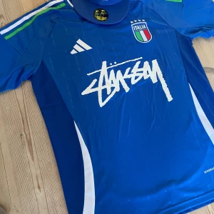 Blå Italien fotbollströja Adidas x Stüssy - Snygg blå fotbollströja från Adidas i samarbete med Stüssy och Italien. Tröjan har korta ärmar, vita och gröna detaljer på axlarna, samt Italiens landslagsemblem på bröstet. Materialet är lätt och ventilerande Aeroready. Stüssy-logga i vitt framtill.