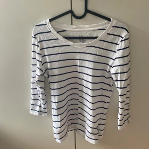 Randig vit topp med knappar - Vit topp med marinblå ränder från H&M. Trekvartsärm med snygga knappar vid ärmslutet. Mjuk och skön bomullskvalitet, perfekt för vår och sommar. Enkel och stilren design som passar till det mesta.