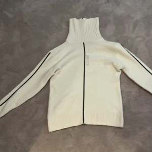 Vit polotröja Boss med svarta detaljer - Vit polotröja  med högt ribbat krage och svarta linjer längs ärmar och mitt fram. Tröjan har lång ärm, ribbade muddar och glittrig text. 