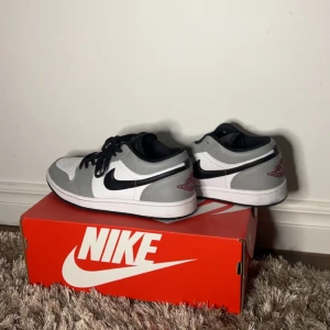 Nike Jordan 1 Low Grå/Vit US 8.5 - Nike Jordan 1 Low sneakers i grått, vitt och svart med klassisk svart swoosh på sidan. Skorna har snörning, perforerad tå och röd logga på hälen. Låg modell med platt sula och rund tå. Tillverkade i skinn och syntetmaterial. Perfekt för dig som gillar stilrena sneakers.