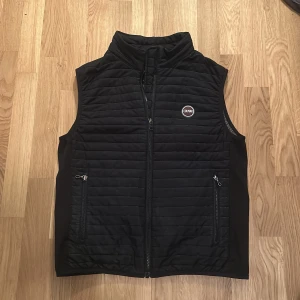 Svart Colmar väst  - Svart dunväst från Colmar med quiltad design och hög krage. Västen har två praktiska fickor med dragkedja framtill och ett Colmar-märke på bröstet. Perfekt för lager-på-lager och snygg till streetwear. Stängs med dragkedja framtill.