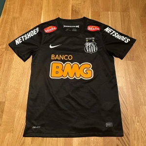 Svart Santos Neymar Jr fotbollströja Nike - Säljer en svart Santos fotbollströja med Neymar Jr och nummer 11 på ryggen. Tröjan har korta ärmar, Dri-Fit material och flera sponsorer som BMG, Netshoes och Seara. Klubbmärket är tryckt på bröstet och Nike-loggan syns tydligt. Perfekt för dig som gillar brasiliansk fotboll.
