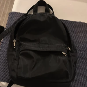Svart ryggsäck från Forever 21 - En stilren svart ryggsäck från Forever 21 i syntetmaterial. Väskan har ett stort huvudfack, en praktisk ytterficka med dragkedja och justerbara axelremmar. Perfekt för skolan eller stan när du vill ha med dig allt du behöver.lite mindre än vanlig ryggsäck 