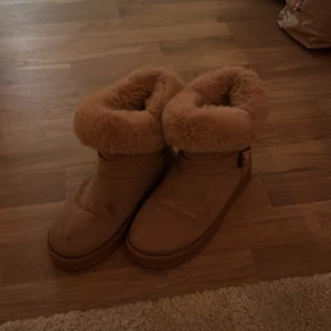 Beiga boots med fluffig päls - Mysiga boots i beige med rund tå och platt sula. Skorna har en bred, fluffig pälskant upptill som ger extra värme och en chill vinterlook. Utsidan är i mocka och insidan är fodrad med mjuk päls. Perfekta för kalla dagar när du vill vara både varm och snygg.