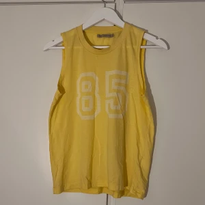Gult linne från NLY Trend - Gult linne från NLY Trend i storlek XS. Toppen har siffran 85 tryckt framtill och rund halsringning. Ser väldigt gul ut på bilderna, men i verkligeheten är färgen mycket mildare och ljusare. 