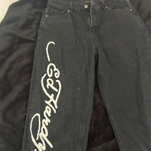 Svarta Ed Hardy jeans med dödskalletryck - Svarta jeans från Ed Hardy med stort vitt logotryck längs ena benet och feta dödskallemotiv med eldflammor på bakfickorna. Jeansen har en loose passform och raka ben, tillverkade i klassisk denim. Perfekta för dig som vill sticka ut med edgy streetstyle. Använda max 3 gånger. Köptes för 1300kr. Säljer dem för att de inte är min stil längre och någon annan kan ta nytta av dem. Köpte dem i en större storlek då de är perfekt för en loose-fit. Pris går att diskuteras.