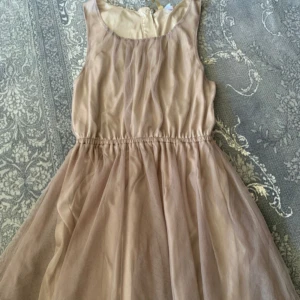 Beige ärmlös klänning med tunn tyll - Supersöt beige klänning med tunn tyll och markerad midja. Den är ärmlös och har en rund halsringning samt dragkedja bak. Perfekt för dig som gillar en romantisk och luftig stil.