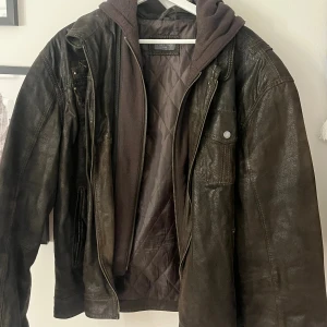 Mörkbrun skinnjacka med huva från just top  - Säljer en mörkbrun skinnjacka med avtagbar huva i sweatshirtmaterial. Jackan har flera fickor med tryckknappar och dragkedja framtill. Quiltat innerfoder ger extra värme och en cool look. Perfekt för dig som gillar streetstyle och vill ha en jacka med attityd.