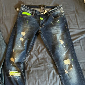 Philipp Plein skinny jeans blå 31 - Snygga skinny jeans från Philipp Plein i mörkblå denim med slitningar, färgstänk i grönt och vitt samt coola neonfärgade detaljer. Klassisk femficksmodell med unika trasiga partier och grönt märke bak. Perfekta för dig som vill sticka ut. 