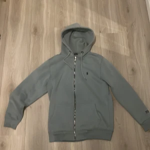 Grå hoodie från Polo Ralph Lauren - Står Medium men sitter och känns som Large.(Därför jag säljer den eftersom trodde den skulle vara Medium men den passa inte) Snygg grå hoodie från Polo Ralph Lauren med dragkedja, huva med snörning och broderad logga på bröstet. Mjuk insida och två fickor framtill. Klassisk och stilren design med Polo-brodyr även på ärmen.