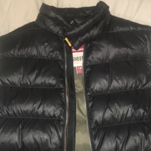 Svart pufferjacka från Parajumpers - Säljer en svart pufferjacka från Parajumpers, modell Sheen. Jackan har quiltad design, hög krage och dragkedja framtill. Insidan är olivgrön och jackan har en cool orange detalj i nacken. Perfekt för kalla dagar och riktigt snygg streetstil.