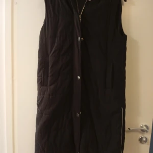 Svart lång dunväst från Boohoo - Svart quiltad dunväst från Boohoo i storlek M. Västen är lång, har huva, praktiska fickor och dragkedja längs sidan. Stängs med både dragkedja och tryckknappar framtill. Perfekt för lager-på-lager under kyliga dagar.