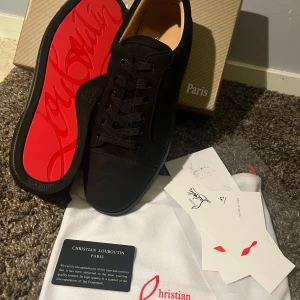 Christian Louboutin svarta sneakers - Svarta sneakers från Christian Louboutin med lyxig mocka och ikonisk röd sula med logotyp. Skorna har rund tå, platt sula och snörning framtill. Diskret broderad logga på hälen och mjuk innersida i beige. Perfekt för dig som vill sticka ut med exklusiv stil.