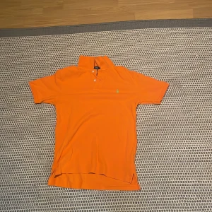 Orange pikétröja från Polo Ralph Lauren - Snygg orange pikétröja från Polo Ralph Lauren med klassisk krage och två knappar framtill. Tröjan har korta ärmar och den ikoniska gröna loggan broderad på bröstet. Perfekt för en fräsch och sportig look.