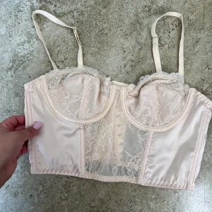 Champagnefärgad spetskorsett - Säljer en champagnefärgad korsett med justerbara axelband och vackra spetsdetaljer framtill och på kuporna. Toppen har hakknäppning bak och är tillverkad i satin och spets för en lyxig känsla. Perfekt för dig som vill ha en feminin och trendig look.