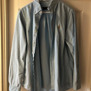 Ralph Laurent skjorta  - Snygg ljusblå skjorta från Polo Ralph Lauren i bomull med klassisk button-down krage och broderad logga på bröstet. Skjortan har lång ärm, vita knappar och en stilren, normal passform. Perfekt för dig som gillar clean och tidlös stil.