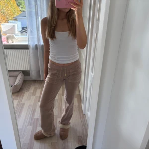 Beiga mjukisbyxor med snörning - Säljer ett par beiga mjukisbyxor i mjuk velour med snörning i midjan och guldiga detaljer. Byxorna har raka ben, fickor fram och bak samt en liten broderad logga på sidan. Perfekta för en chill dag hemma.