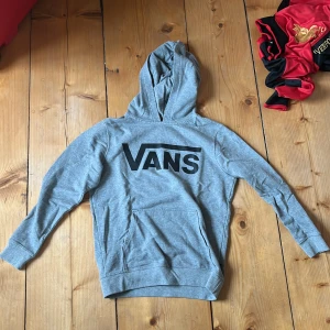 Grå hoodie från Vans med logga - Grå hoodie från Vans i storlek medium med stor svart Vans-logga på bröstet och texten 'Off The Wall' på insidan av huvan. Tröjan har klassisk känguruficka, ribbade muddar och är tillverkad i mjuk bomullsmix. Perfekt för chill dagar och streetstyle.