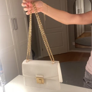 Vit axelväska med guldkedja - Snygg vit axelväska med stilren design och guldfärgad kedja som axelrem. Väskan har ett elegant guldfärgat spänne framtill och är tillverkad i ett slätt, strukturerat material. Perfekt för att lyfta vilken outfit som helst. Köpt på new yorker