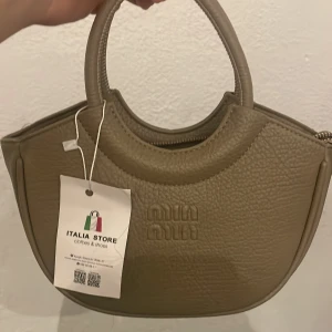 Beige handväska från Miu Miu - Snygg beige handväska från Miu Miu i mjukt skinn med rundade handtag och guldfärgad dragkedja. Väskan har en stilren, halvmåneformad design och kommer med en matchande axelrem. Perfekt för dig som gillar minimalistisk och trendig look.