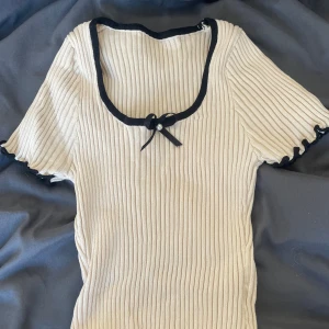 Beige ribbad topp med rosett - Supersöt croppad topp i beige ribbat material med svarta detaljer längs kanterna och en liten rosett med pärla vid urringningen. Toppen har korta ärmar med vågig kant och sitter tight. Perfekt för dig som gillar en söt och trendig stil.