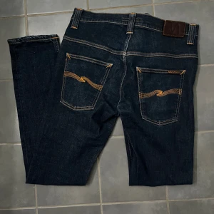 Mörkblå Nudie Jeans Tape Ted W32 L34 - Snygga mörkblå jeans från Nudie Jeans, modellen Tape Ted. Klassisk femficksdesign med orange kontrastsömmar och vågig brodering på bakfickorna. Tillverkade i 98% ekologisk bomull och 2% elastan för skön stretch. Perfekt passform och stilren look. Hör av dig vid minsta fråga!