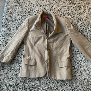 Elegant beige Hugo Boss kavaj (Storlek M) - Snabb leverans. Stilren och modern kavaj från Hugo Boss, perfekt för både vardag och mer formella tillfällen. Den är i beige färg, har en snygg passform och kommer i storlek M. Jackan är lätt och bekväm, med två fickor på framsidan. Ett utmärkt val för att lägga till en sofistikerad touch till din garderob. Perfekt för den stilmedvetna!