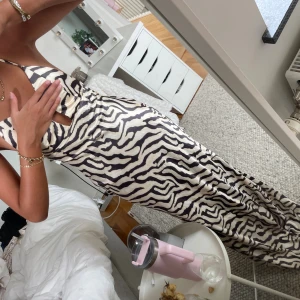 Zebramönstrad långklänning med öppen rygg - Säljer en långklänning med zebramönster i svart och beige. Klänningen har djup v-ringning, tunna axelband och öppen rygg med snygg snörning. Materialet känns glansigt och svalt, perfekt för sommaren. Sitter figurnära och ger en riktigt trendig look. Aldrig använd!