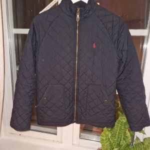 Mörkblå quiltad jacka från Ralph Lauren - Quiltad mörkblå jacka från Ralph Lauren med guldiga dragkedjedetaljer och klassisk röd logga på bröstet. Jackan har två fickor framtill med tryckknappar och hög krage. Perfekt för höst och vår, stilren och tidlös design.rivan i fickan syns inte när man har på den bara om du öppnar fickan och kolla efter den och i vuxen storlek är det s om du undrar varför det står så i den 