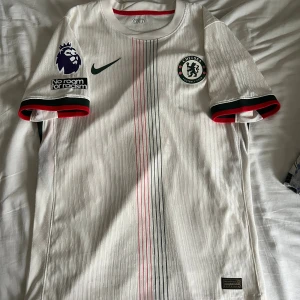Chelsea FC bortatröja 25/26 - Chelsea FC:s eleganta bortatröja för säsongen 2025/26 – vit med tunna röda och gröna ränder framtill. Officiell Nike Dri-Fit ADV med Premier League-armbadge och ”No Room For Racism”-märke. Mycket bekväm och snygg design – perfekt både till matchdag och vardag.  ✅ Storlek: M ✅ Passform: Normal ✅ Skick: Bra, små skavanker ✅ Märke: Nike (Dri-Fit ADV)  📦 Skickas snabbt och välpaketerad.