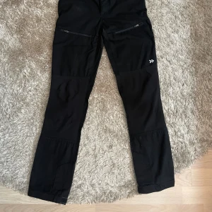 Svarta täckbyxor från Funk, strl 160 - Svarta täckbyxor från Funk med raka ben och praktiska dragkedjor på framsidan. Byxorna har bälteshällor och förstärkta partier vid knäna. Perfekta för utomhusaktiviteter under kyliga dagar. Snygg och funktionell design med diskret logga på benet.