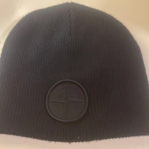 Svart mössa från Stone Island - Säljer en svart ribbstickad mössa från Stone Island med klassisk rund logotyp framtill i svart ton-i-ton. Mössan är tillverkad i mjukt material och har en clean, stilren look som passar perfekt till streetwear och vinteroutfits.