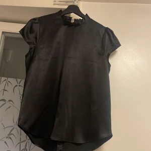 Svart blus med volang från River Island - Snygg svart blus från River Island i glansigt material med korta puffärmar och volangdetalj runt halsen. Blusen har en liten öppning med knapp i nacken, perfekt för dig som gillar stilrena och trendiga plagg.