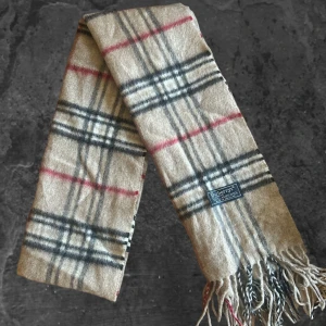 halsduk från Burberry - Klassisk rutig halsduk från Burberrys i beige, svart, rött och vitt. Tillverkad i 100% lammull och har fransar i ändarna. Den ikoniska rutiga designen gör den till ett riktigt statement-plagg för vintern.