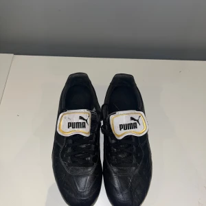 Svarta Puma fotbollsskor i skinn - Klassiska svarta fotbollsskor från Puma med vita detaljer och guldkanter på plösen. Skorna har snörning, rund tå och dobbar för bra grepp på planen. Tillverkade i skinn för en schysst känsla och passform. Perfekta för dig som vill ha stilrena och funktionella fotbollsskor.