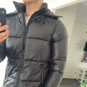 Svart pufferjacka från BoohooMAN - Säljer en svart pufferjacka från BoohooMAN med huva och dragkedja. Jackan är vadderad och har en glansig finish, perfekt för kalla dagar. Snygg och enkel design med två fickor framtill och hög krage.