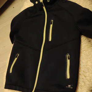 Svart vindjacka från Zara Boys - Svart vindjacka från Zara Boys med gula dragkedjor och detaljer. Jackan har huva, tre fickor med dragkedja och meshfoder i gult. Perfekt för blåsiga dagar och snygg streetstil. Inga anmärkningar. Nyskick men har använts. 