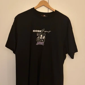 Svart Kobe Bryant Lakers t-shirt (Black Mamba) - Svart t-shirt från BRO med tryck av Kobe Bryant, nummer 24 och Los Angeles Lakers-logga både fram och bak. Texten 'BLACK MAMBA' på ryggen och coola motiv i lila och vitt. Klassisk passform och rund hals. Perfekt för basketfans!