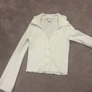 Vit blus med krage från Monki - Säljer en vit blus från Monki i storlek XXS. Blusen har en krage med lätt volangkant, v-ringning och knappar framtill. Materialet är strukturerad polyester med lite stretch. Långärmad med lite utsvängd ärm och lätt croppad passform, perfekt för en clean och fräsch look.